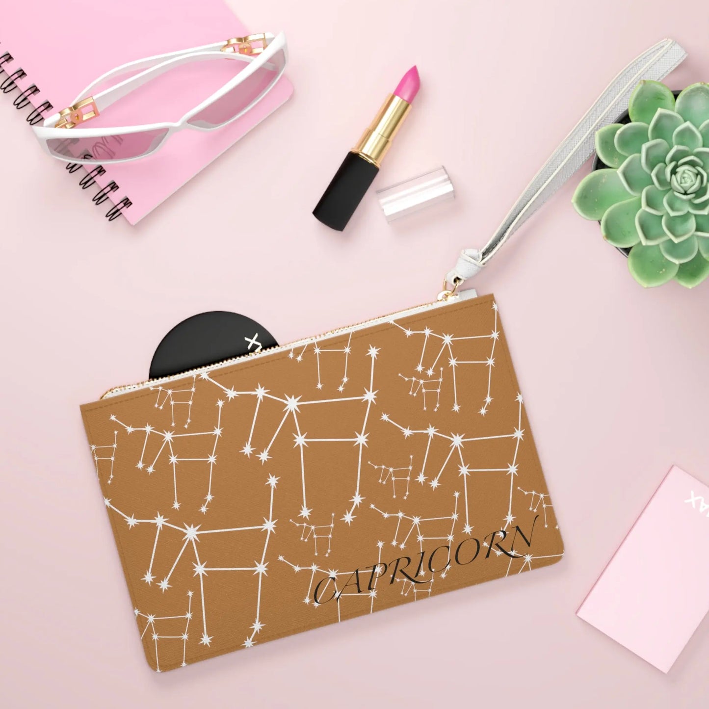 Capricorn Zodiac Clutch shown in a lifestyle setting with sunglasses and lipstick on a pink background.