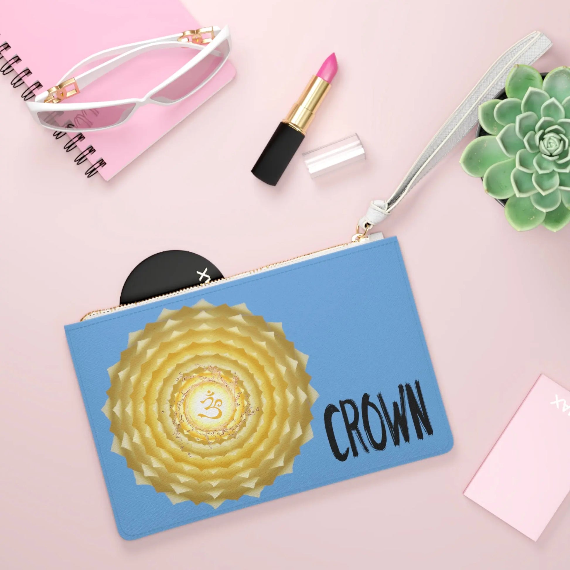 Crown Chakra Clutch shown in a lifestyle setting with sunglasses and lipstick on a pink background.