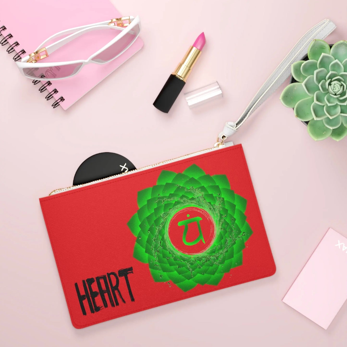 Heart Chakra Clutch shown in a lifestyle setting with sunglasses and lipstick on a pink background.