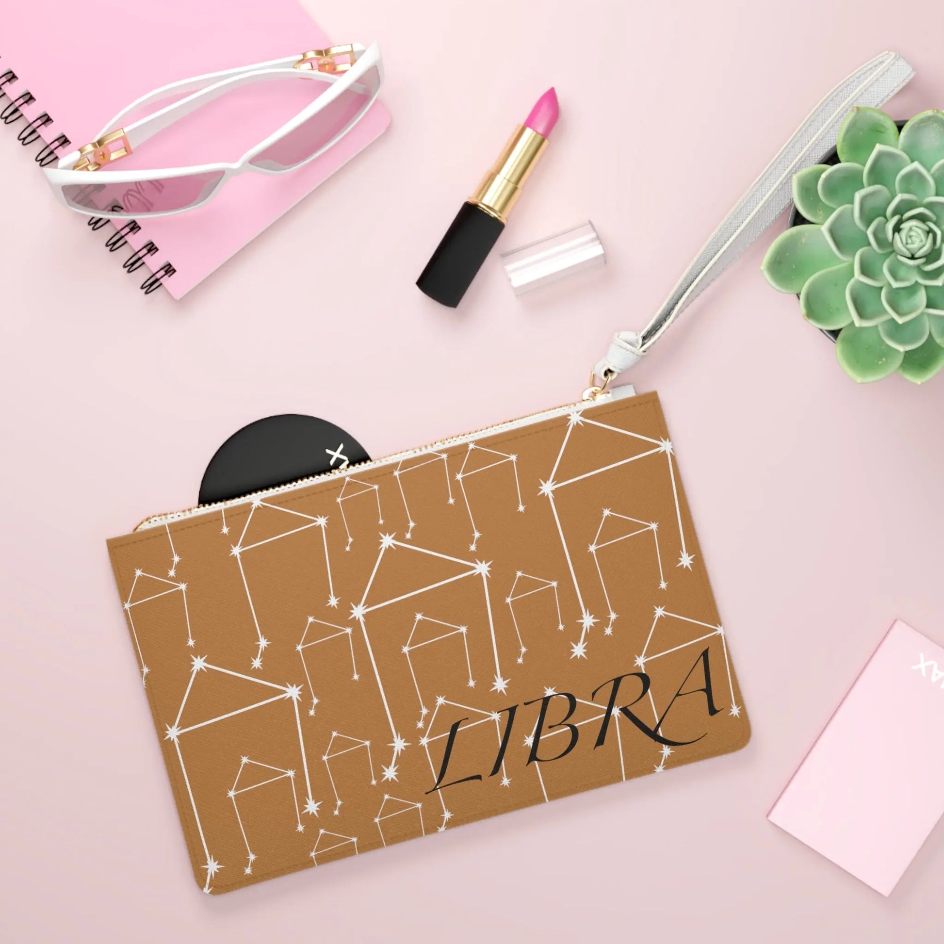 Libra Zodiac Clutch shown in a lifestyle setting with sunglasses and lipstick on a pink background.