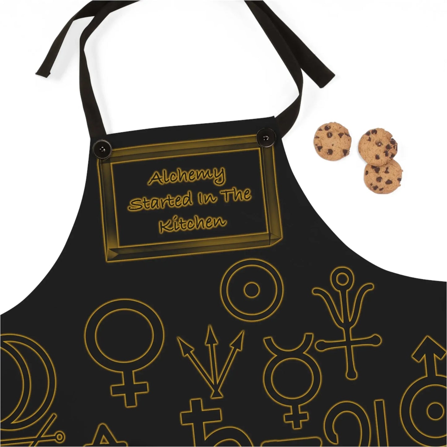 Black apron with 'Alchemy Started In The Kitchen' text and gold astrological symbols, paired with chocolate chip cookies.