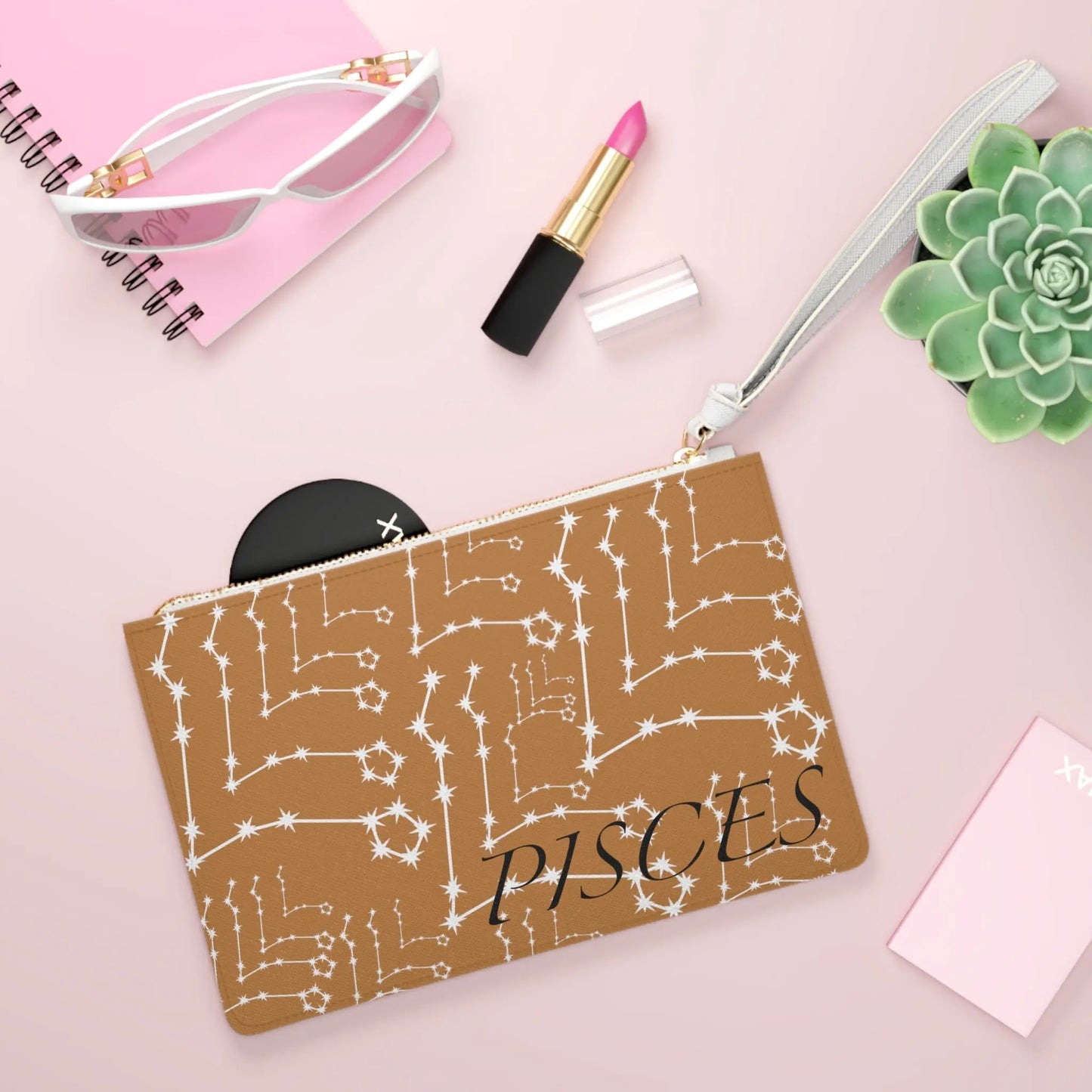 Pisces Zodiac Clutch shown in a lifestyle setting with sunglasses and lipstick on a pink background.