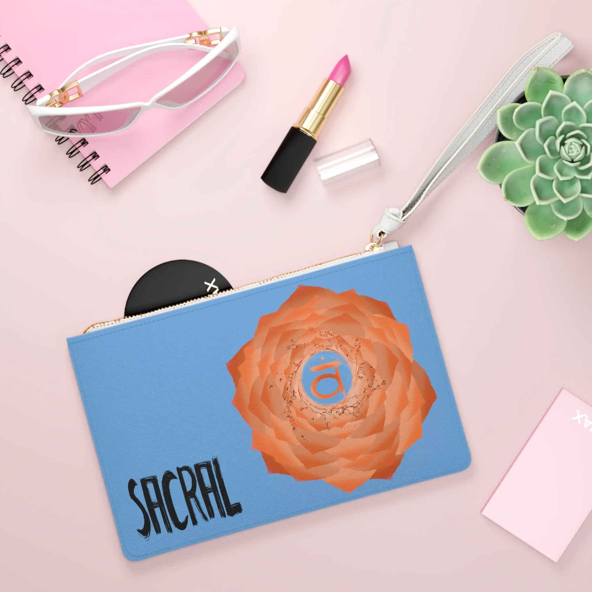 Sacral Chakra Clutch shown in a lifestyle setting with sunglasses and lipstick on a pink background.