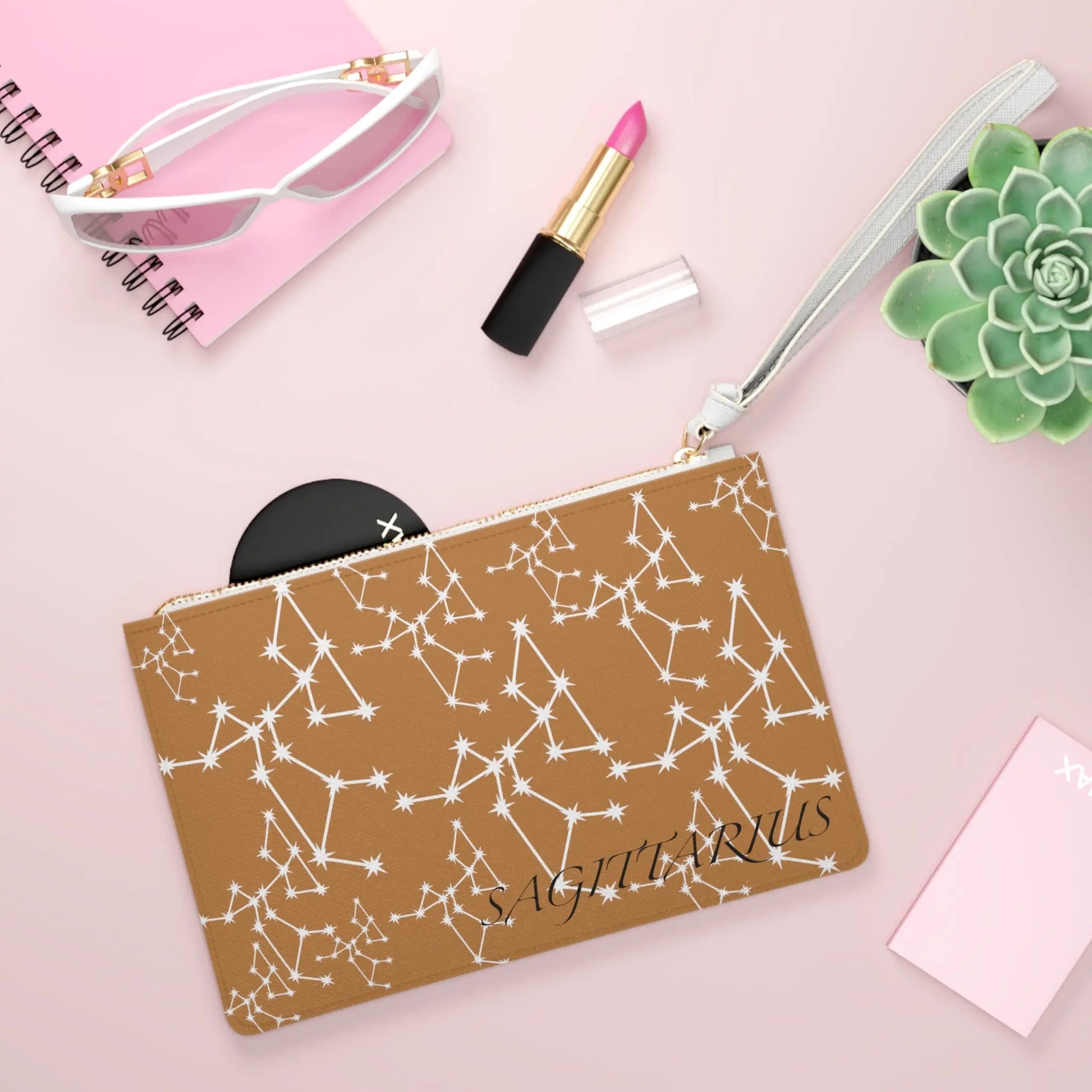 Sagittarius Zodiac Clutch shown in a lifestyle setting with sunglasses and lipstick on a pink background.