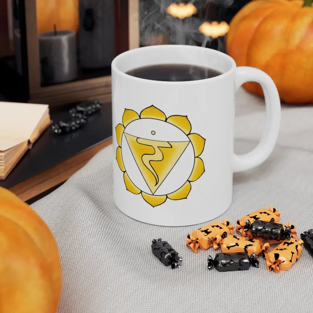 White mug with yellow chakra symbol, steaming coffee, Halloween candies, and pumpkins for spiritual autumn vibes