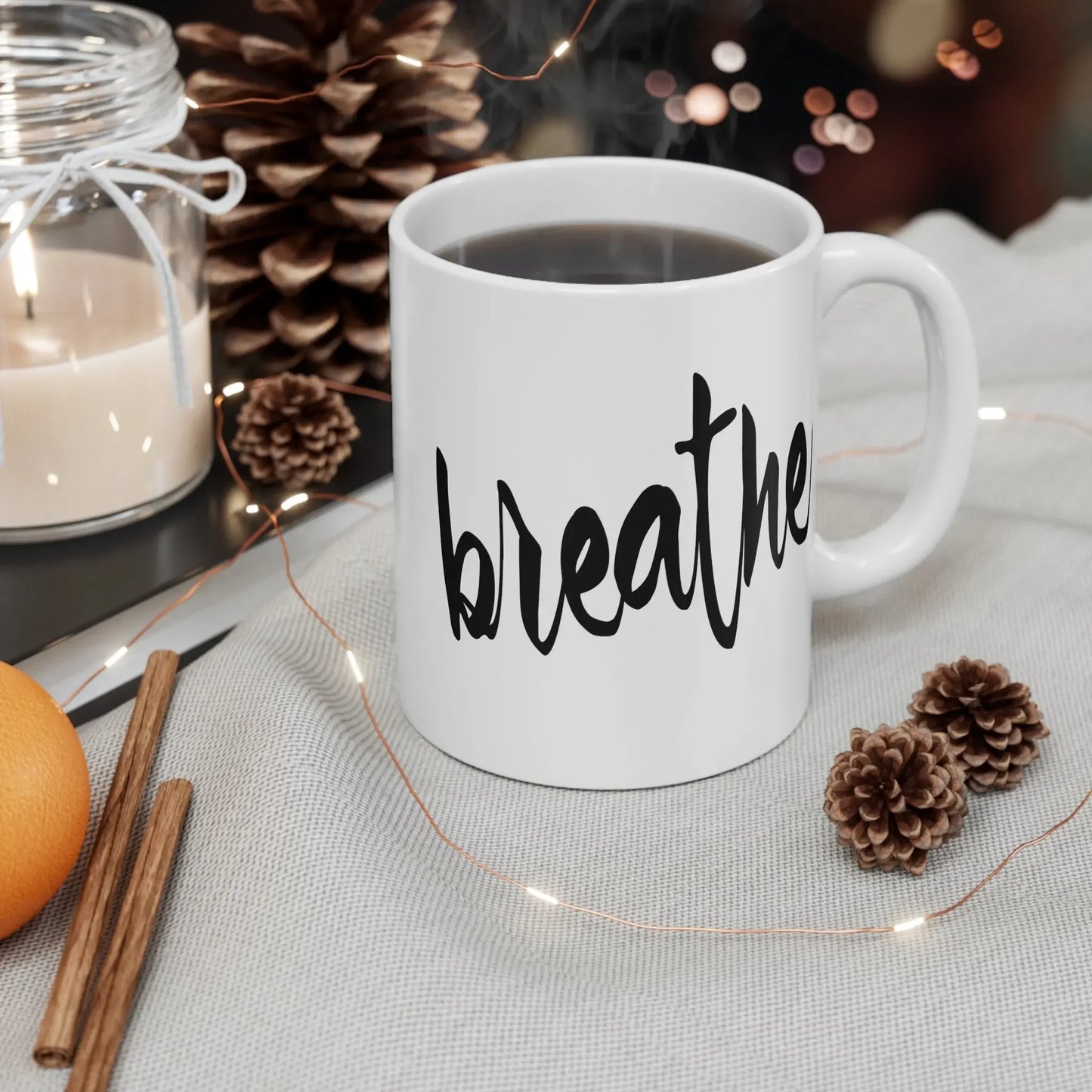 White ceramic mug with 'breathe' text, steaming coffee, pinecones, candle, and fairy lights for mindful moments