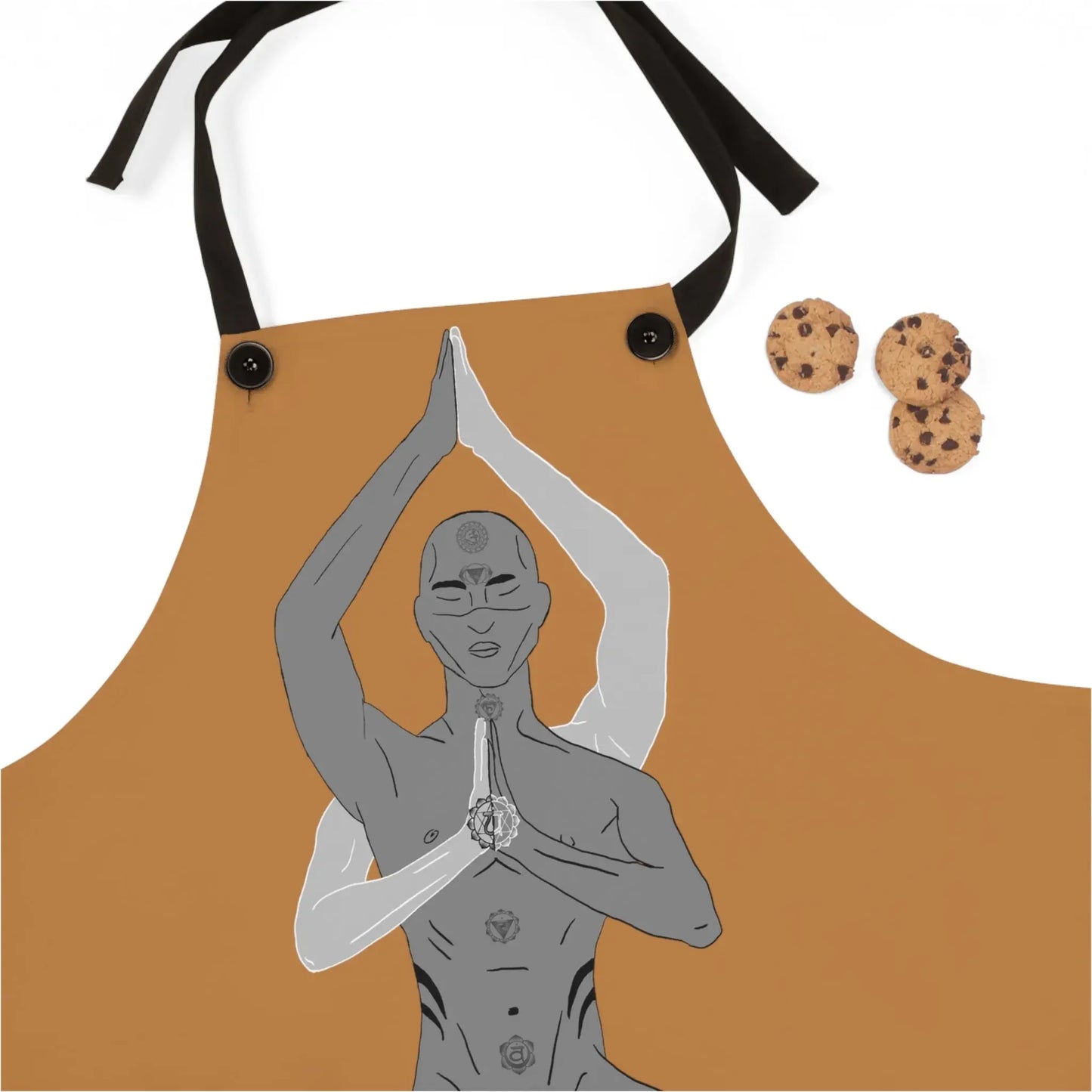 Tan apron with chakra yoga figure design, black straps, and chocolate chip cookies nearby