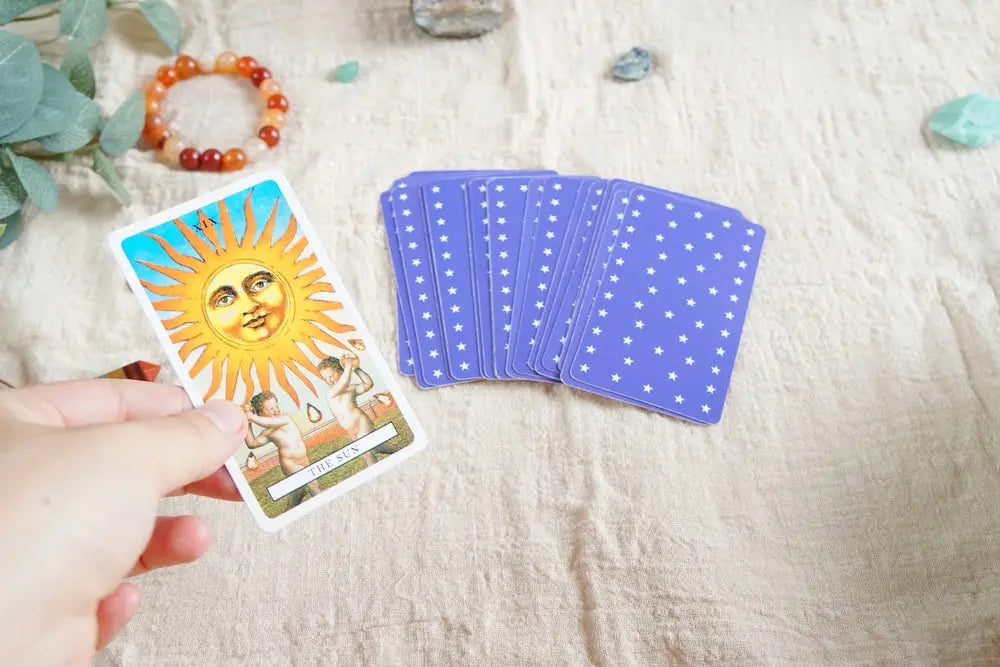 The Sun tarot card with purple star-patterned cards, crystals, and a beaded bracelet on fabric.